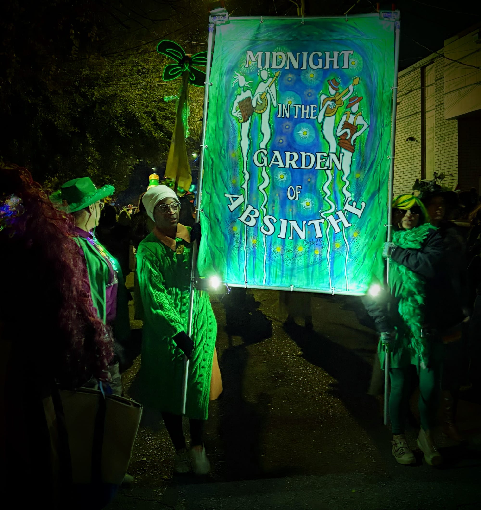 two greenclad women holding a painted sign reading MIDNIGHT IN THE GARDEN OF ABSINTHE